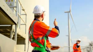 Two workers in orange safety gear and helmets stand near wind turbines. One gestures thumbs up, creating a collaborative and positive atmosphere.